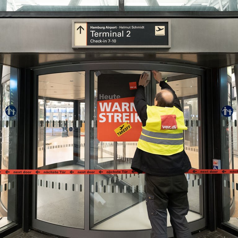 Ein Verdi-Vertreter hängt ein Plakat mit der Aufschrift «Warnstreik» an einer geschlossenen Terminaldrehtür am Hamburger Flughafen auf.